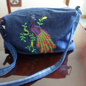 May include: A blue denim crossbody bag with an embroidered peacock design. The peacock is in shades of purple, green, yellow, and red.