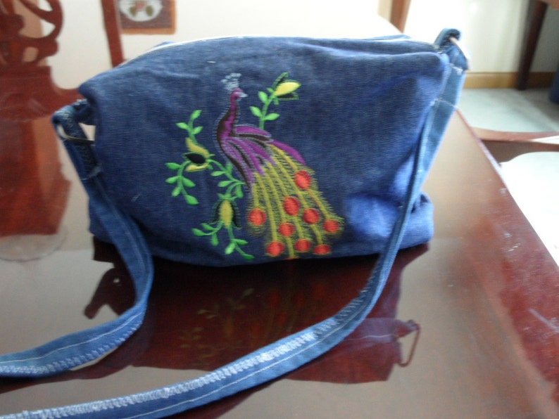 May include: A blue denim crossbody bag with an embroidered peacock design. The peacock is in shades of purple, green, yellow, and red.