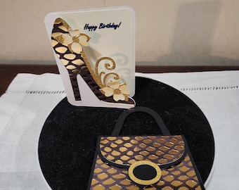 Op de afbeelding: Een wenskaart met een ontwerp van een high-heelschoen en de tekst "Happy Birthday!". De schoen is bruin en goudkleurig met bloemaccenten. Daaronder staat een bijpassende handtas met een goud- en bruin patroon.
