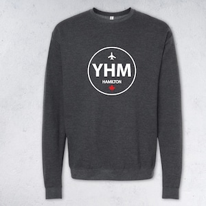 May include: Dark grey sweatshirt featuring a white circular graphic. The graphic includes the letters "YHM", an airplane icon, the word "Hamilton", and a red maple leaf. The sweatshirt has a crew neck.