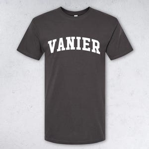May include: Dark grey t-shirt featuring the white word "VANIER" in a bold, arched font. The short-sleeved shirt is shown against a light grey background.