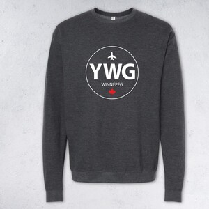 Peut inclure: Sweat-shirt gris foncé avec un motif circulaire blanc. Le motif comprend les lettres "YWG", le mot "Winnipeg", un petit avion et une feuille d'érable rouge. Le sweat-shirt a un col rond et des manches longues.
