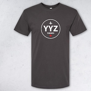 May include: Dark grey t-shirt with a white circular graphic. The graphic includes the letters "YYZ", a small airplane, and the word "Toronto" with a red maple leaf. The shirt has a crew neck.