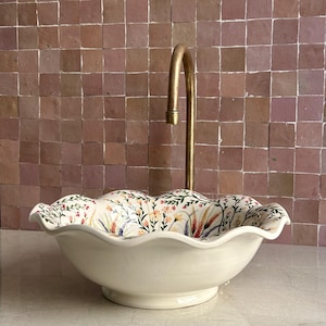May include: Cream-colored ceramic sink with a floral pattern on the rim, featuring a gold-toned faucet. The sink has a scalloped edge and sits on a countertop against a pink tile wall. A decorative bathroom fixture.