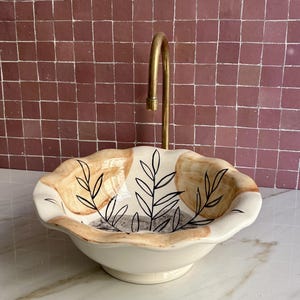 Peut inclure: Un évier en céramique avec un robinet en laiton. L'évier blanc cassé a un bord festonné et un motif peint à la main de feuilles noires et de formes beiges. Le robinet a un bec courbé. Idéal pour une salle de bain.