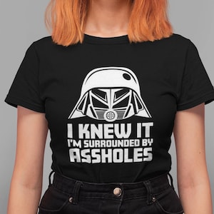 May include: Black t-shirt with a white graphic of a helmet and the text "I KNEW IT I'M SURROUNDED BY ASSHOLES". The shirt is a crew neck and has short sleeves. The model is wearing black jeans.