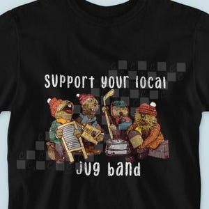 May include: Black t-shirt featuring a cartoon band of four characters playing musical instruments. The text "Support your local jug band" is printed in white above the band. The band members are wearing hats and playing a washboard, a jug, and other instruments.