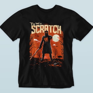 May include: Black t-shirt featuring a graphic of a knight with a sword, standing in front of a red and orange sunset with the text "'Tis but a SCRATCH". The design includes a full moon and bare trees.