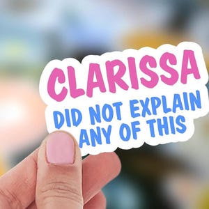 May include: A white sticker with the text "CLARISSA DID NOT EXPLAIN ANY OF THIS" in pink and blue. The sticker is held by a person with a blurred background.