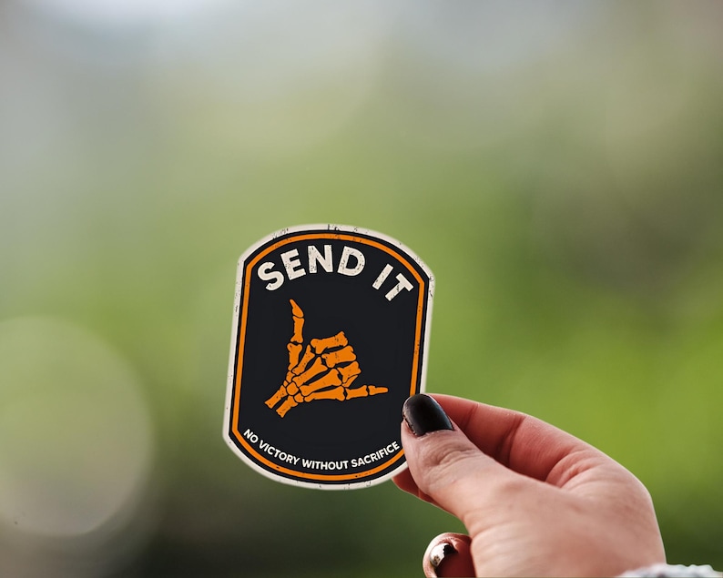 May include: A black and orange sticker with the words "SEND IT" and a skeleton hand making a shaka sign. The sticker also includes the text "NO VICTORY WITHOUT SACRIFICE".