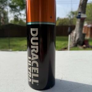 Pode incluir: Um copo cil&iacute;ndrico com um design que imita uma bateria Duracell Ultra. A se&ccedil;&atilde;o superior &eacute; laranja, a do meio &eacute; preta e a inferior tem o logotipo Duracell Ultra em branco. O copo &eacute; feito de a&ccedil;o inoxid&aacute;vel e foi projetado para manter as bebidas frias.