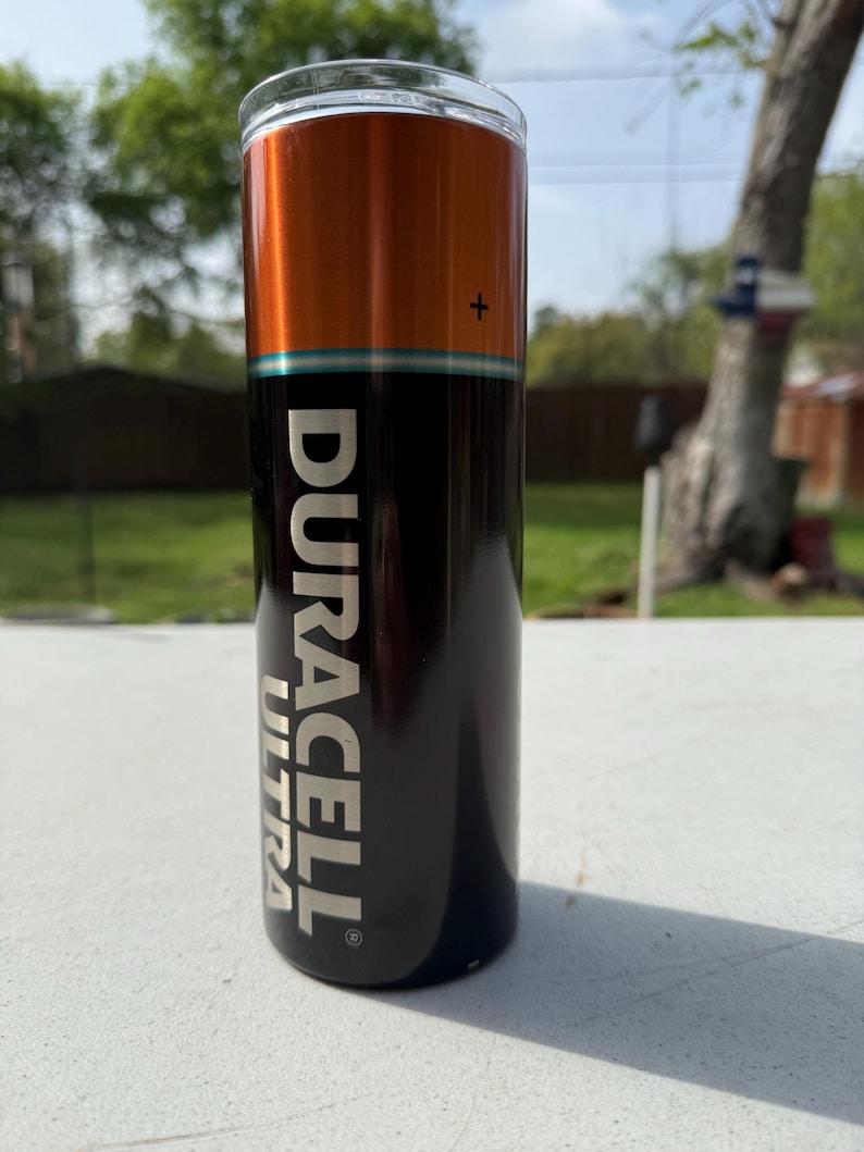 Pode incluir: Um copo cil&iacute;ndrico com um design que imita uma bateria Duracell Ultra. A se&ccedil;&atilde;o superior &eacute; laranja, a do meio &eacute; preta e a inferior tem o logotipo Duracell Ultra em branco. O copo &eacute; feito de a&ccedil;o inoxid&aacute;vel e foi projetado para manter as bebidas frias.