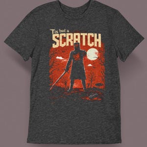 May include: Dark gray t-shirt featuring a graphic of a knight holding a sword, with the text "'Tis but a Scratch" in white. The design includes a red background with a full moon and trees.