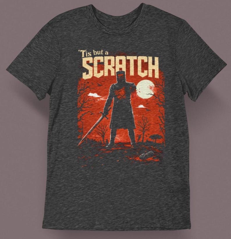 May include: Dark gray t-shirt featuring a graphic of a knight holding a sword, with the text "'Tis but a Scratch" in white. The design includes a red background with a full moon and trees.