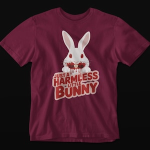 May include: A maroon t-shirt featuring a white bunny with blood around its mouth. The text "JUST A HARMLESS LITTLE BUNNY" is printed in red and white below the bunny graphic. The shirt is made of a soft material.
