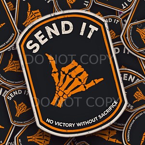 May include: Black sticker with the words "SEND IT" in white, featuring an orange skeleton hand in the shaka gesture. The phrase "NO VICTORY WITHOUT SACRIFICE" is printed below the hand.