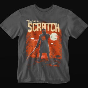May include: Dark gray t-shirt featuring a graphic of a knight with a sword, standing in front of a red and orange background with a full moon. The text "'Tis but a SCRATCH" is above the knight.