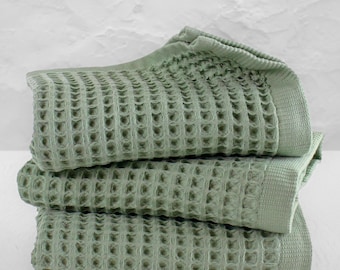 May include: A stack of three sage green towels featuring a distinct waffle weave texture. Each towel has a smooth, flat-woven border along the edge, showcasing a consistent light green color throughout the folded fabric layers.