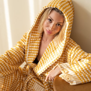 May include: A hooded bathrobe featuring a textured, waffle-like pattern in alternating mustard yellow and white stripes. The robe includes long sleeves with fringed cuffs and a matching tie belt at the waist, crafted from a soft, absorbent fabric.