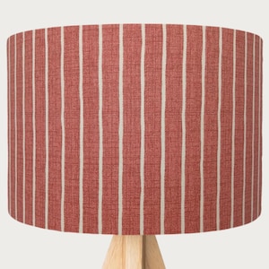 Puede incluir: Pantalla de lámpara cilíndrica con un patrón de rayas rojas y blancas. La pantalla está hecha de una tela texturizada y se asienta sobre una base de madera clara. Las rayas verticales están espaciadas uniformemente.