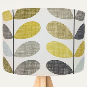 Peut inclure: Abat-jour cylindrique avec un motif de feuilles dans des tons de jaune, gris et vert olive sur fond blanc. Il repose sur un socle en bois clair, créant un design moderne et inspiré de la nature.