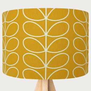 May include: Mustard yellow lampshade featuring a white leaf design. The cylindrical lampshade sits on a light-coloured wooden base. The leaf pattern is a repeating design of stylised, oval-shaped leaves.
