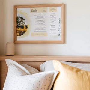 Peut inclure: Un cadre en bois clair contenant une impression avec le mot allemand "Erde" et du texte, avec une image de paysage d'un arbre et de tournesols. Le cadre est monté sur un mur blanc au-dessus d'un lit avec des oreillers.