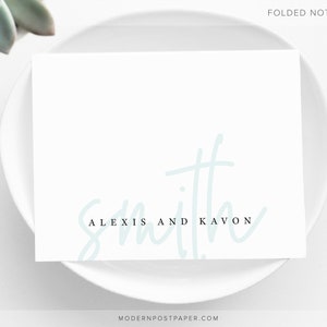 May include: A white folded note card with the text "Alexis and Kavon Smith" in black and a light blue cursive font.