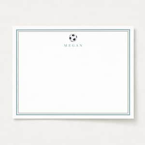 May include: A rectangular white stationery card featuring a thin double-line blue border. Centered at the top is a small black and white soccer ball graphic positioned above the name MEGAN printed in a simple blue sans-serif font.