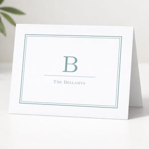 May include: A white folded note card featuring a blue double-line border, a large blue letter B centered at the top, and the text The Bellamys printed in blue below the letter. The card is positioned horizontally on a flat surface.