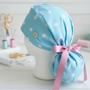 May include: A light blue scrub cap with a white star pattern, a button detail, and a pink ribbon. The cap is displayed on a white mannequin head. Sewing supplies are in the background.