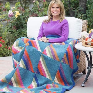 May include: A woman sits outdoors, draped in a vibrant, geometric-patterned blanket. The blanket features a blue base with colorful triangular sections of orange, pink, green, and white stripes. The woman wears a purple sweater.