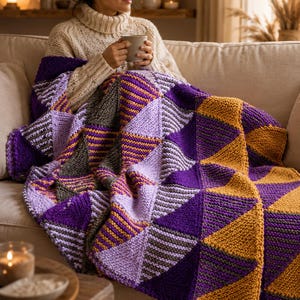 Puede incluir: Una manta de punto con un diseño geométrico de triángulos en tonos morado, dorado y gris. La manta está sobre un sofá, creando un ambiente cálido. Hecha de hilo suave y grueso, ideal para relajarse.