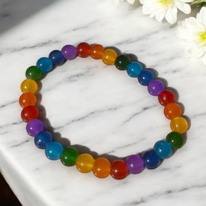 This image features a vibrant rainbow glass bead bracelet displayed on a white marble surface. The bracelet is composed of smooth, glossy round beads arranged in a repeating rainbow pattern, including red, orange, yellow, green, blue, and purple tones. Each bead has a rich, translucent finish that shines beautifully in the light.