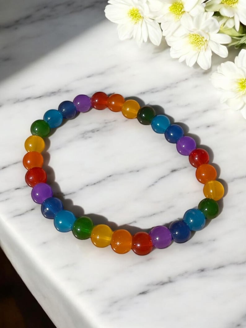 This image features a vibrant rainbow glass bead bracelet displayed on a white marble surface. The bracelet is composed of smooth, glossy round beads arranged in a repeating rainbow pattern, including red, orange, yellow, green, blue, and purple tones. Each bead has a rich, translucent finish that shines beautifully in the light.