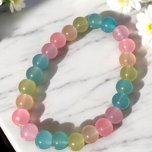 his image showcases a pastel multicolor glass bead bracelet displayed on a white marble surface with soft gray veining. The bracelet is made of smooth, round glass beads in soft, translucent shades of teal, baby pink, peach, yellow-green, and light blue. Each bead has a gentle shine that catches the light, giving the bracelet a luminous and dreamy appearance.