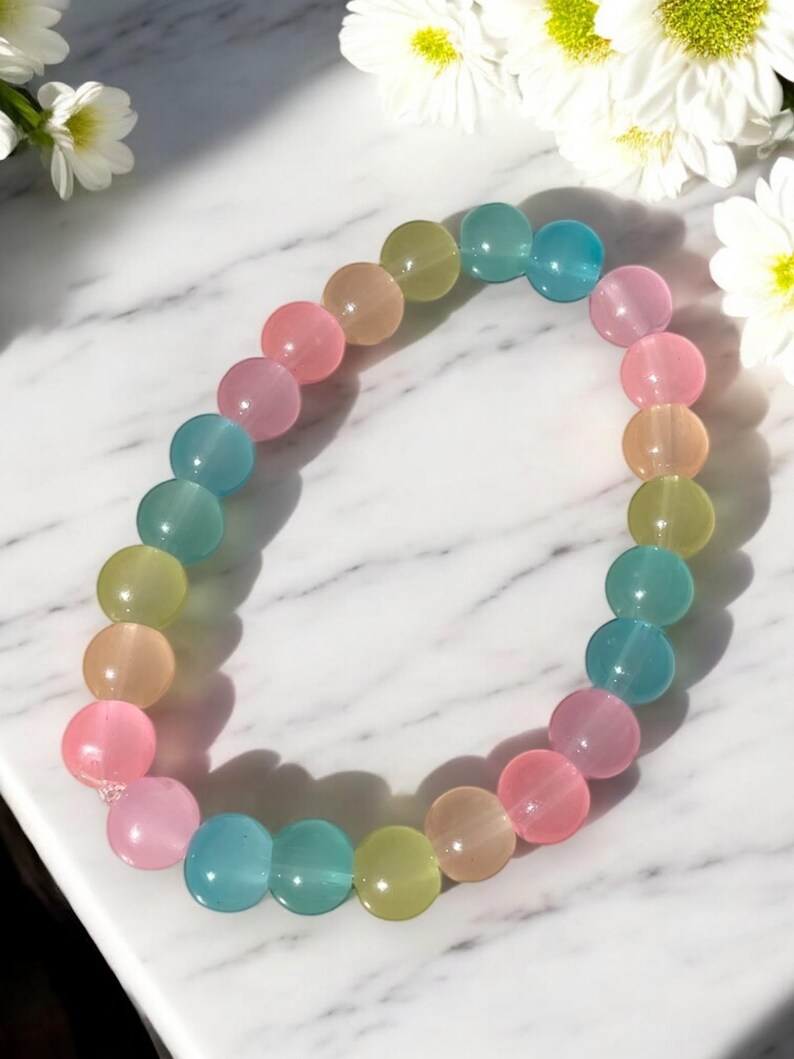 his image showcases a pastel multicolor glass bead bracelet displayed on a white marble surface with soft gray veining. The bracelet is made of smooth, round glass beads in soft, translucent shades of teal, baby pink, peach, yellow-green, and light blue. Each bead has a gentle shine that catches the light, giving the bracelet a luminous and dreamy appearance.