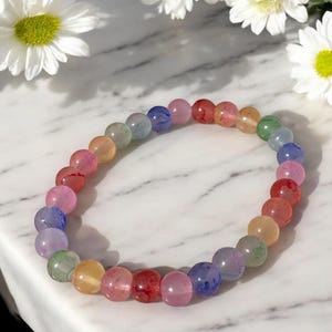 This image features a single handcrafted beaded bracelet resting on a white marble surface. The bracelet is made from round, glossy glass beads in a variety of soft, translucent colors including pink, lavender, red, green, yellow, and blue. Some beads have delicate marbling or veining within them, adding texture and visual interest.