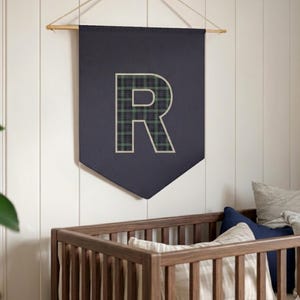 Op de afbeelding: Marineblauwe wandbanner met een geruit "R"-ontwerp. De letter is groen, zwart en wit. De banner hangt boven een houten wieg met kussens.