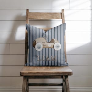 May include: A decorative pillow with a blue and gray striped background. The pillow features a watercolor illustration of a vintage car in shades of brown and beige. The name "LIAM" is printed in brown letters below the car.