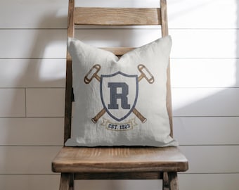 Op de afbeelding: Een decoratief kussen met een marineblauw schildontwerp met de letter "R", gekruiste hamers en de tekst "EST. 1923". Het kussen is off-white en ligt op een houten stoel tegen een witte wand met lambrisering.