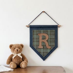 May include: A navy blue and plaid fabric banner with a wooden dowel and a leather letter "R". A brown teddy bear with a white bow sits on a wooden surface. The banner is hanging on a white wall.