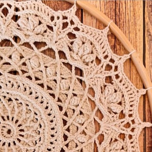 Peut inclure: Attrape-r&ecirc;ves en crochet fait &agrave; la main, de couleur cr&egrave;me, avec des motifs ajour&eacute;s complexes. Le design circulaire est suspendu dans un cerceau en bois naturel. L'artisanat est pr&eacute;sent&eacute; sur un fond en bois.