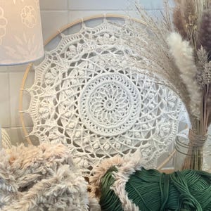 Peut inclure: Un attrape-r&ecirc;ves mandala en crochet cr&egrave;me est pr&eacute;sent&eacute; sur un cerceau en bois. Le motif complexe est centr&eacute; sur un motif en spirale. Une lampe avec un abat-jour floral, de la laine et des fleurs s&eacute;ch&eacute;es dans un bocal en verre sont &eacute;galement dans l'image.