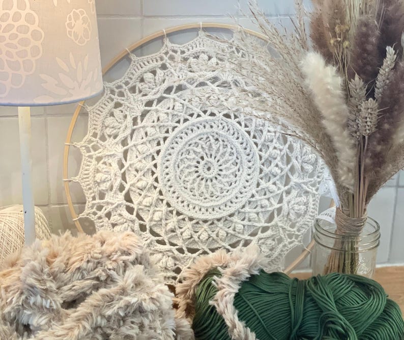 Peut inclure: Un attrape-r&ecirc;ves mandala en crochet cr&egrave;me est pr&eacute;sent&eacute; sur un cerceau en bois. Le motif complexe est centr&eacute; sur un motif en spirale. Une lampe avec un abat-jour floral, de la laine et des fleurs s&eacute;ch&eacute;es dans un bocal en verre sont &eacute;galement dans l'image.