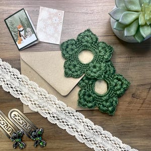May include: An assortment of craft supplies on a wooden surface. Includes two green crocheted flower shapes, a cream-colored lace ribbon, two metal bookmarks with green accents, a small card with a fox illustration, and a succulent plant.