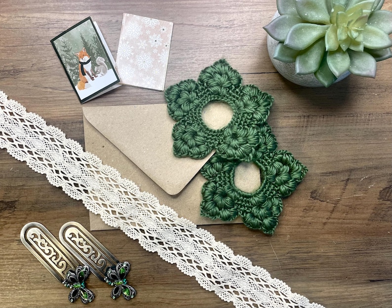 May include: An assortment of craft supplies on a wooden surface. Includes two green crocheted flower shapes, a cream-colored lace ribbon, two metal bookmarks with green accents, a small card with a fox illustration, and a succulent plant.