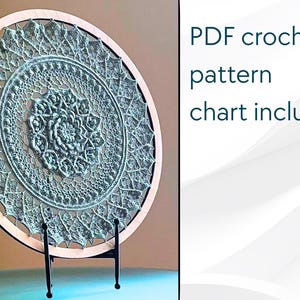 Pode incluir: Um centro de mesa redondo de crochê verde claro exibido em um aro de madeira, montado em um suporte de metal preto. O centro de mesa apresenta um intrincado design floral simétrico. O texto à direita diz: PDF crochet pattern chart included.