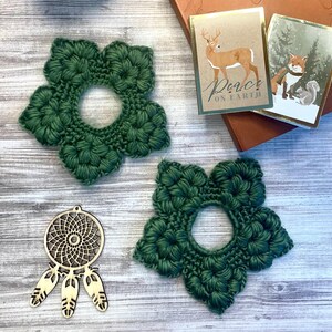 May include: Two dark green, flower-shaped crochet coasters with a central hole are displayed. A wooden dreamcatcher pendant with three feathers and two small holiday cards with gold trim are also visible. The cards feature a deer and a fox.