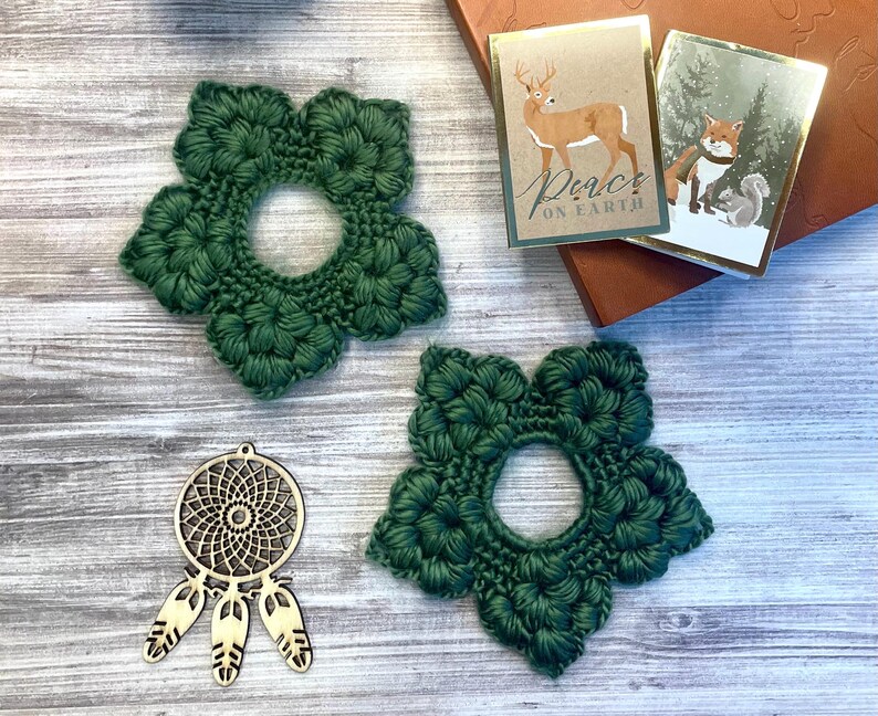 May include: Two dark green, flower-shaped crochet coasters with a central hole are displayed. A wooden dreamcatcher pendant with three feathers and two small holiday cards with gold trim are also visible. The cards feature a deer and a fox.